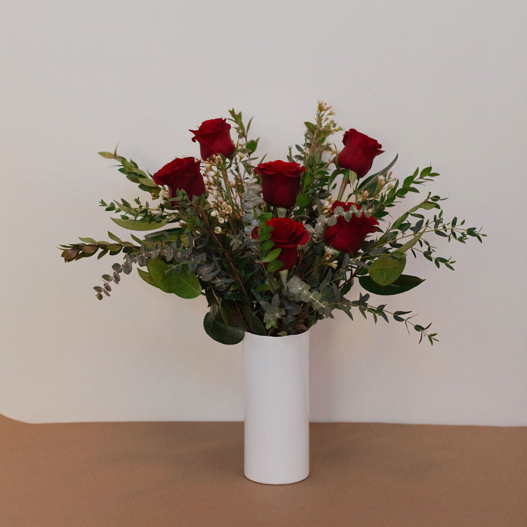 Bouquet of red roses and greenery in a white vase on a beige surface with a light gray background